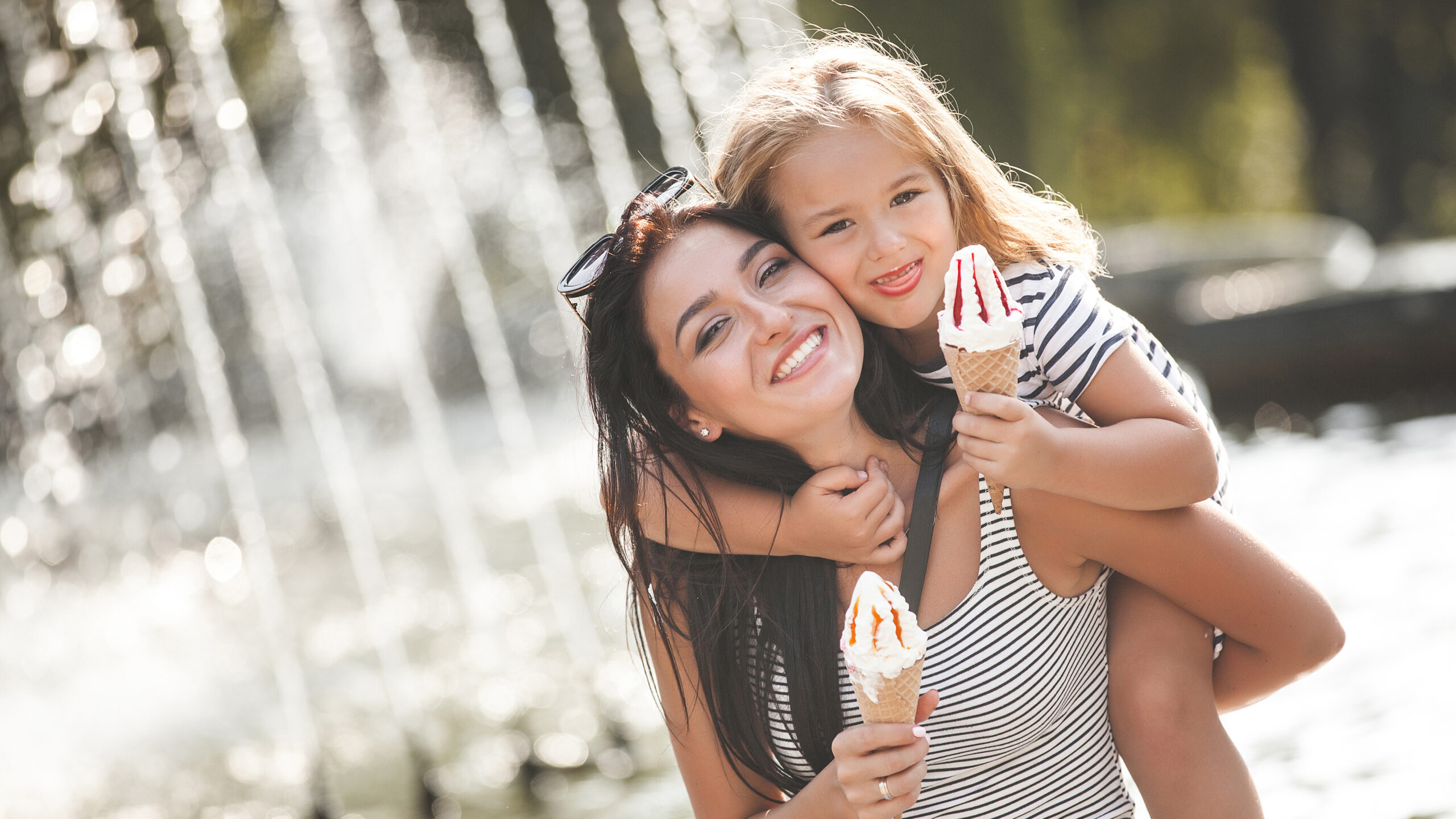 Symbolbild einer Mutter, die mit ihrem Kind vor einem Brunnen fröhlich Eis ist.
